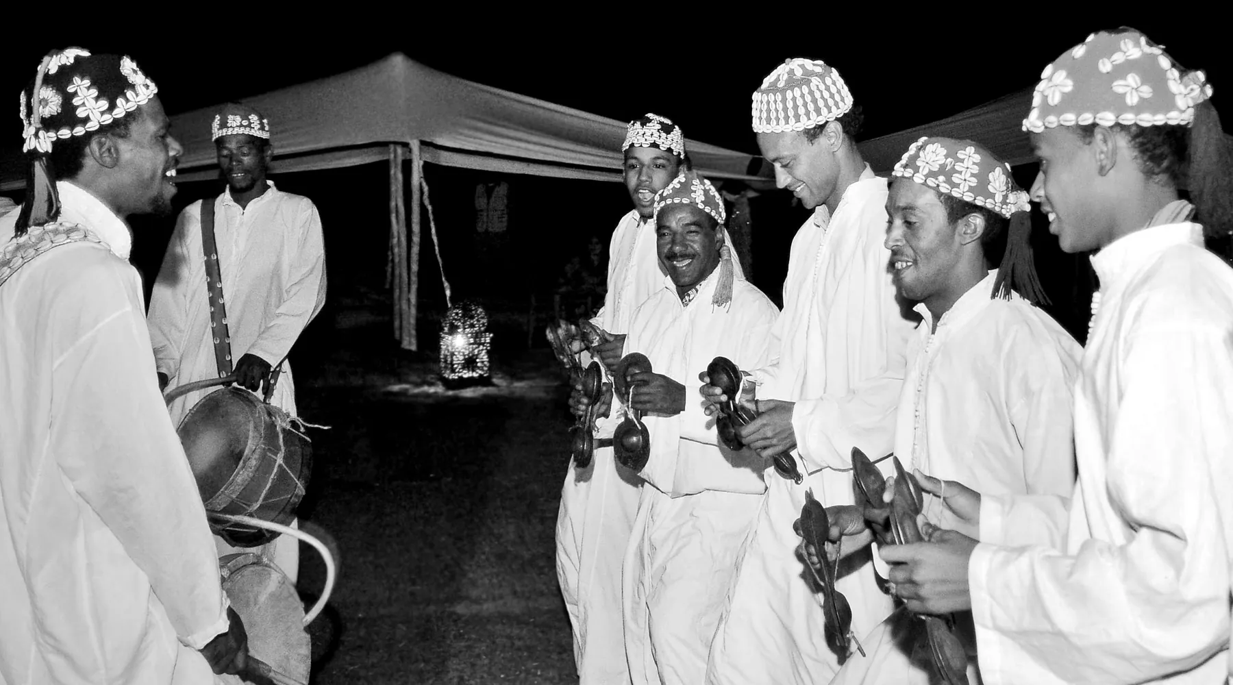 Gnawa musicians in white robes and shell-decorated caps play castanets and a drum outdoors in the Marrakech night
