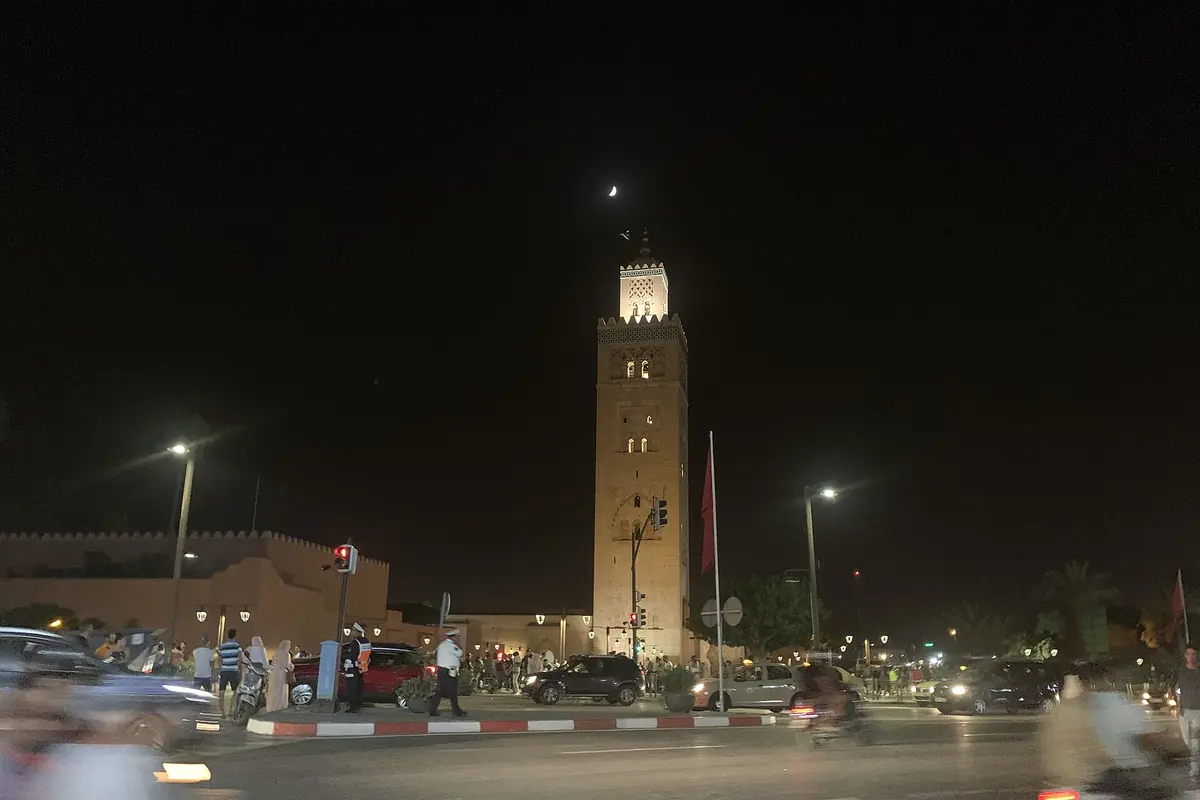 The lit Koutoubia minaret rises above a busy road at night in Marrakech, with blurred traffic in the foreground